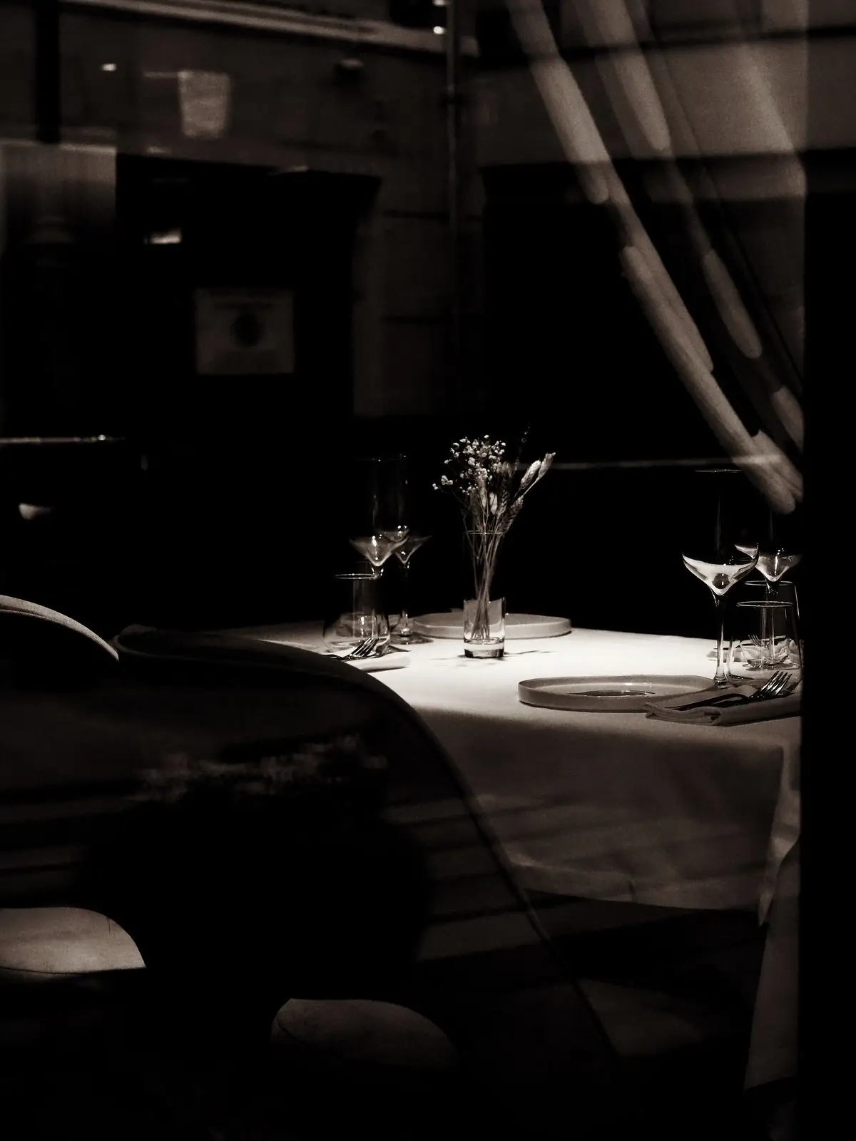 Une table dressée dans la pénombre, vue à travers la vitre, lumière rasante sur la nappe