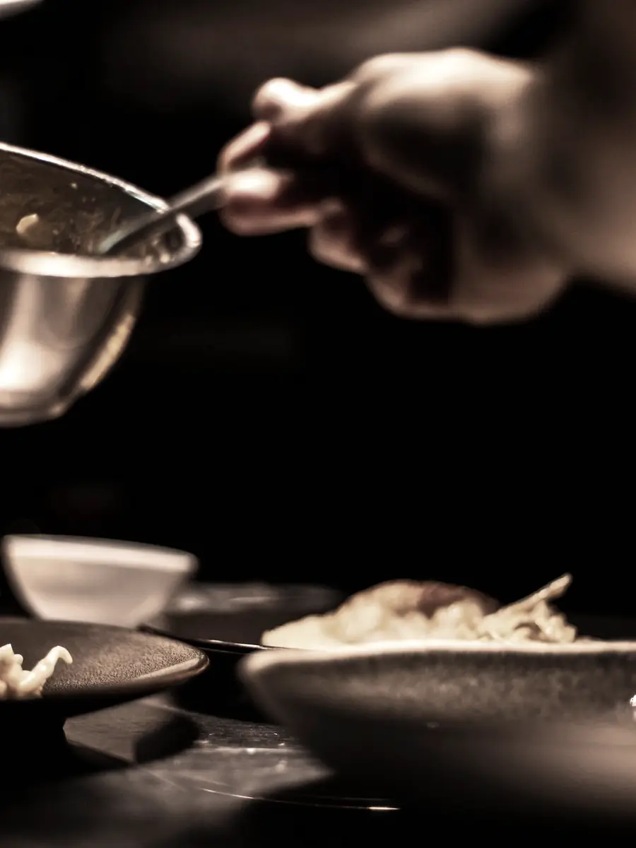 Mains de la cheffe versant au tamis au-dessus d’assiettes, dans la pénombre du passe